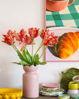 På ett bord med färgglad servis står en rosa vas innehållande Konstblomman Fransk Parakittulpan 76 cm röd/gul, medan i bakgrunden delvis syns en inramad tavla av en croissant och en kopp.