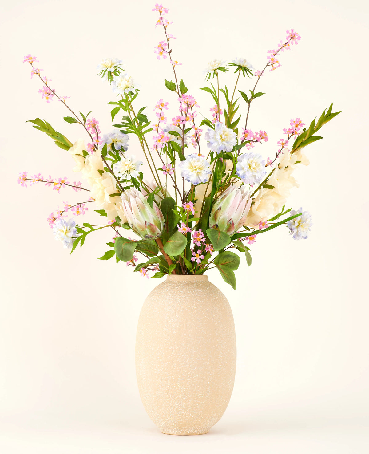 I en vas med beige struktur ligger Silk konstgjorda blombuketten Lots of Love XL, en handbunden bukett av rosa, vita och blå sidenblommor med gröna blad och långa stjälkar mot en ljus bakgrund.