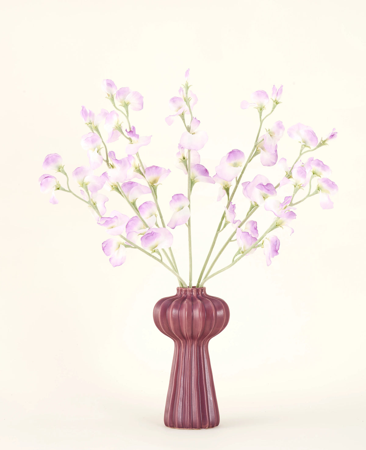 Silk konstgjorda blombuketten Pea lilac har långa stjälkar av ömtåliga, ljuslila blommor och gröna blad i en lila keramisk vas med vertikala åsar, mot en vanlig, benvit bakgrund.