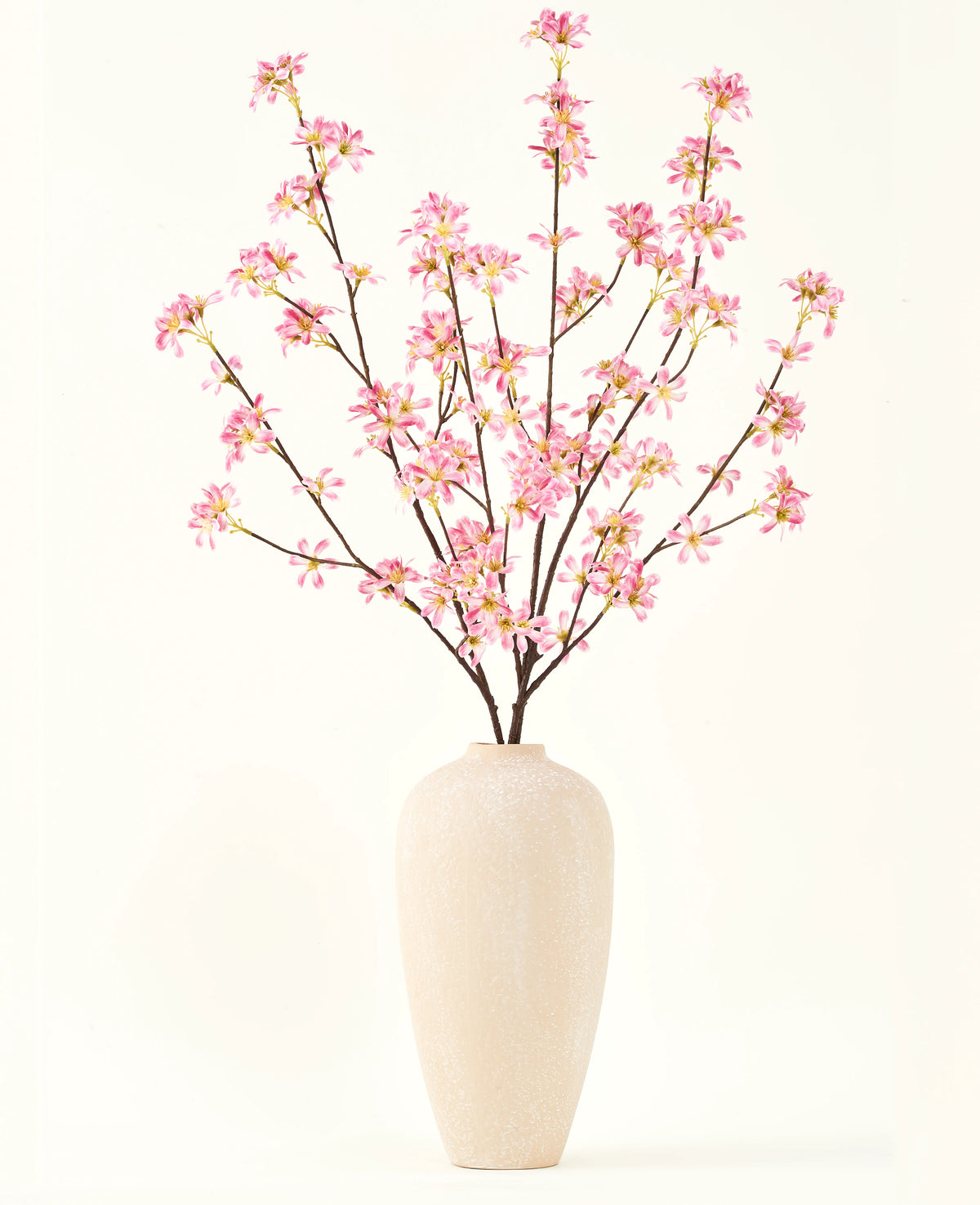 En hög beige keramikvas innehåller flera grenar från Silk konstgjorda blombuketten Rosa blommor, med fina rosa blommor och gröna blad, mot en vanlig vit bakgrund.