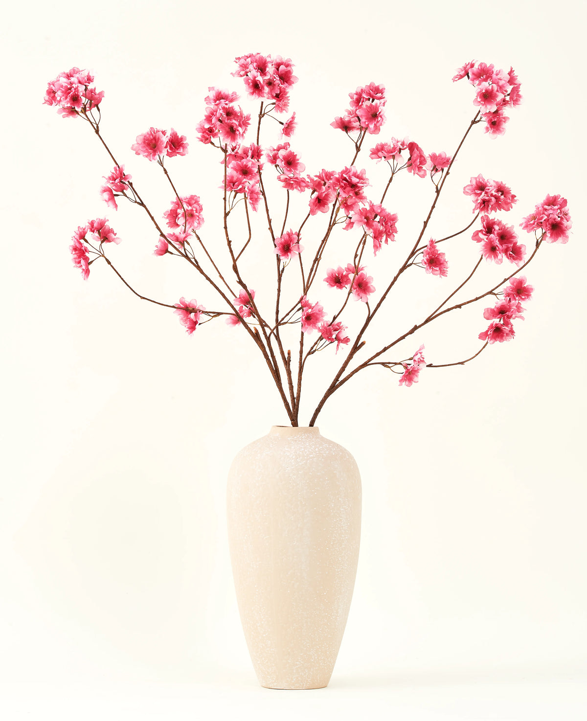 Silk konstgjorda blombuketten Cherry blossom fuchsia 114 cm tillför en livlig färg till din interiör med sina slående grenar och fuchsiablommor, vackert utställda i en beige keramikvas mot en vanlig benvit bakgrund.