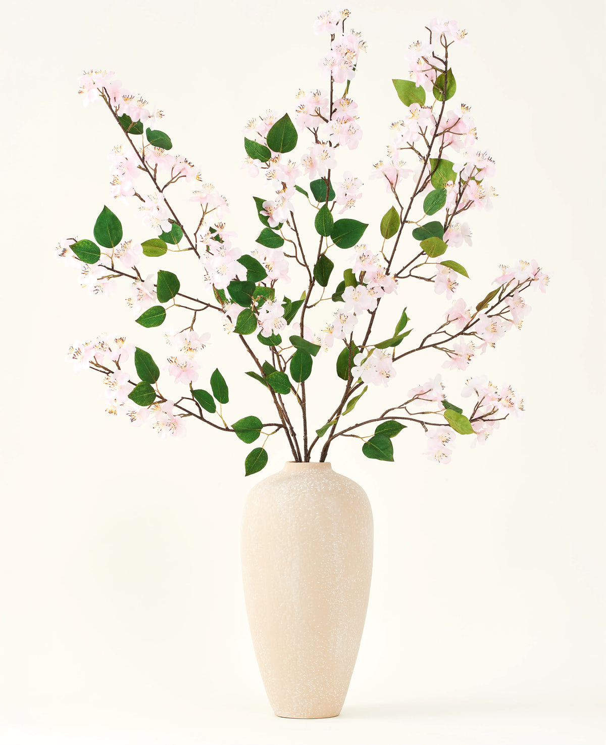 En beige keramikvas innehåller flera långa grenar med Silk artificiell blombukett av Peach Blossom rosa och gröna blad, mot en vanlig vit bakgrund - perfekt för en konstgjord blommig inredning.