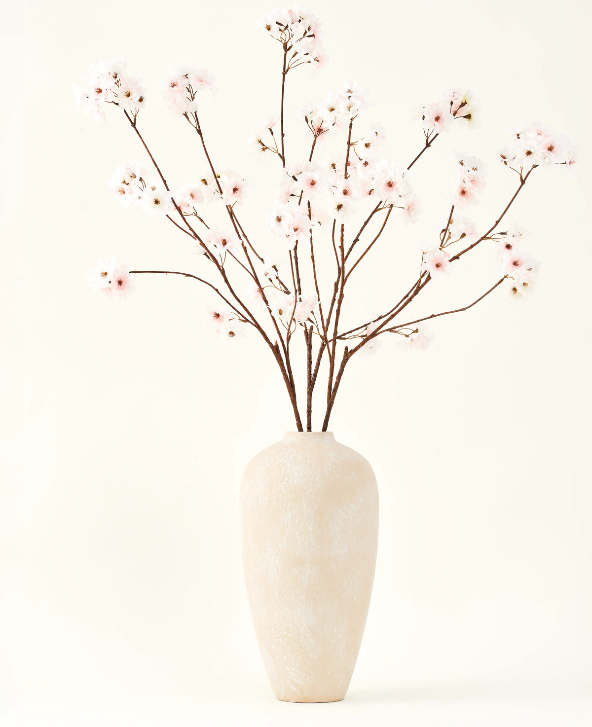 Silk konstgjorda blombuketten Cherry blossom rouge 114 cm har höga, känsliga grenar i en enkel beige vas, som ger en mjuk, elegant touch till din inredning mot en enkel, ljus bakgrund.