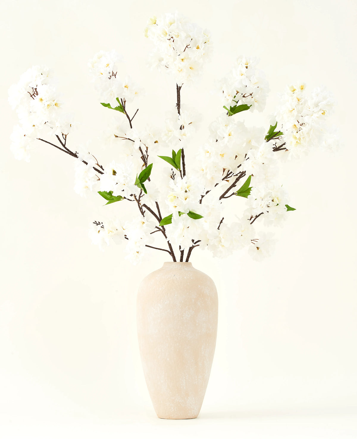Silk konstgjorda blombuketten Cherry blossom cream 119 cm har eleganta bruna grenar med mjuka vita falska blommor och gröna blad, utställd i en enkel beige vas mot en enkel ljus bakgrund.