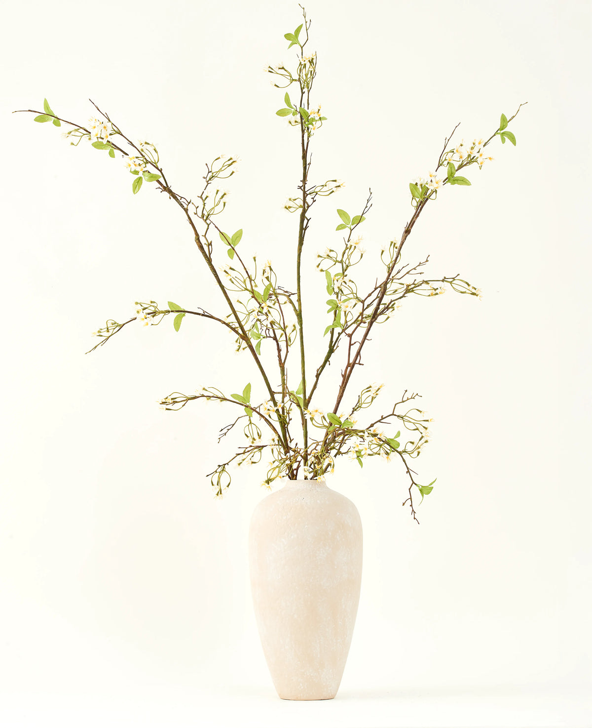 En hög, krämfärgad keramikvas innehåller Silk konstgjorda blombuketten Verbena kräm med långa grenar, små gröna blad och fina vita blommor, vackert arrangerade mot en vanlig ljus bakgrund.
