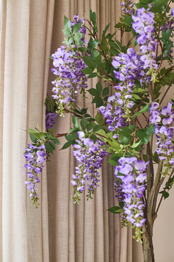 Voor de beige gordijnen hangt een kunstplant Blauweregen van 150 cm met groene bladeren.