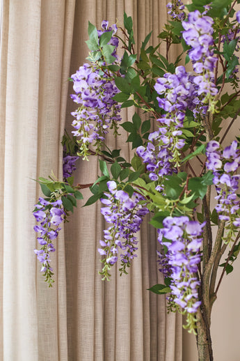 Framför de beiga gardinerna hänger en konstgjord Wisteria-växt på 150 cm med gröna blad.