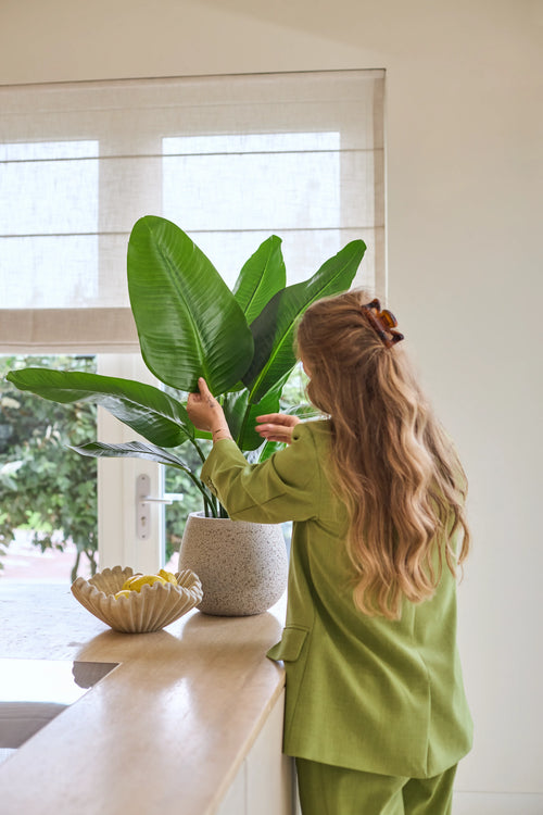 En kvinna med långt vågigt hår, klädd i grön kostym, sköter en 90 cm Strelitzia Real Touch Deluxe konstgjord växt på en disk bredvid en skål med citroner i ett ljust rum med ett fönster.