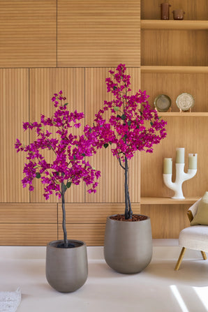 Två krukväxter av typen Artificiell Bougainvillea på 180 cm, i färgen lila, med vibrerande lila blommor står framför en ljus trävägg och hyllor med prydnadsföremål som ljus och tallrikar. Till höger finns en beige stol, som ger en mysig atmosfär till detta livliga konstgjorda växtvardagsrum.