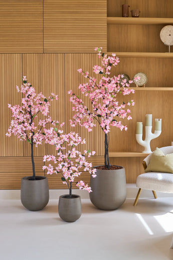 Drie potplanten van het type Kunstplant Bougainvillea van 150 cm, in roze met roze bloemen, staan opgesteld voor houten planken, naast een moderne witte stoel met een kussen en een decoratieve kandelaar.