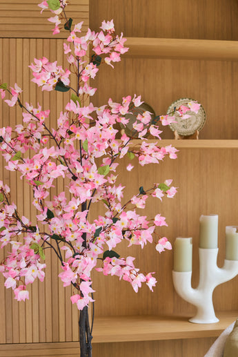 Een kunstplant bougainvillea van 150 cm, roze van kleur en met roze bloesems, staat voor houten planken waarop een decoratief bord en een moderne witte kandelaar met drie groene kaarsen staan uitgestald.