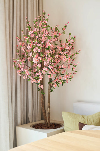 De Kunstplant Kersenbloesem 120 cm roze, met zijn roze kersenbloesembloemen, staat naast een houten tafel in een lichte, moderne kamer, met daarachter een beige gordijn en een lichte bank.