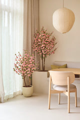Een minimalistische kamer met lichte houten meubels, crèmekleurige gordijnen en twee Kunstplant Kersenbloesem van 150 cm in roze, die voor roze bloesems zorgen. Boven de eettafel, met een stoel met kussens en een ingebouwde bank, hangt een ronde papieren lantaarn.