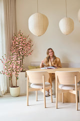 Een vrouw met lang blond haar zit aan een lichte houten tafel, een boek te lezen en glimlachend. Naast haar staat de Kunstplant Kersenbloesem 150 cm roze, waarvan de roze bloesems een levendig accent geven. Hanglampen en een minimalistische inrichting zorgen voor een lichte, gezellige sfeer.