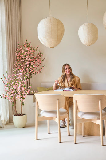 Een vrouw zit glimlachend aan een tafel met een open boek. De kamer is ingericht met lichte houten meubels, papieren lantaarns, zacht natuurlijk licht dat door hoge gordijnen naar binnen valt, en een Kunstplant Kersenbloesem 120 cm roze die een vleugje roze bloesem toevoegt.