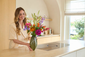 En leende kvinna arrangerar buketten 'Artificial Silk Flowers Colorful Svenski' i en grön vas på en ljus bänkskiva i ett kök med modern inredning och naturligt ljus.
