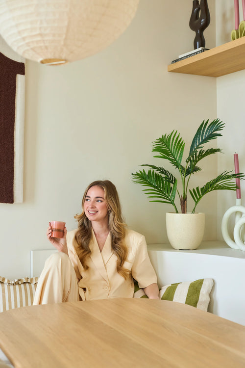 En leende kvinna med långt blont hår sitter i ett modernt, ljust rum med en rosa kopp i handen. Omkring henne finns gröna växter, inklusive den frodiga 85 cm Artificiella Phoenix-växten, samt trähyllor och prydnadsföremål.