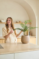 Een vrouw met lang blond haar glimlacht terwijl ze een Kunstpalm Chamaedorea van 48 cm in een pot verzorgt in een moderne, in neutrale tinten gehouden keuken met een minimalistische inrichting.