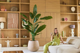 En kvinna i grön outfit sitter i en beige soffa med en drink i handen, med Konstgjorda växten Strelitzia Nicolai Deluxe 210 cm bredvid sig och trähyllor med prydnadsföremål i bakgrunden.