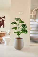 Den 120 cm konstgjorda växten Monstera är placerad i ett modernt, minimalistiskt vardagsrum med neutrala väggar, en rund spegel, trämöbler och abstrakt väggkonst.