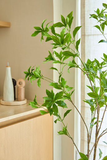 En 150 cm konstgjord Pieris Japonica-växt står vid ett fönster bredvid ett ljust träskåp, som rymmer keramiska flaskor och en pepparkvarn, vilket ger en minimalistisk touch till det ljusa, luftiga rummet.