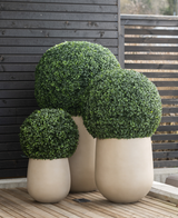 Drie grote Kunstplant Buxus romeinse stijl UV D55cm vormsnoeistruiken met dicht groen gebladerte staan in hoge, beige plantenbakken op een houten terras voor een donker hekwerk met latten.