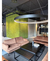 En modern sittgrupp med beige soffor, svarta soffbord, en stor rund taklampa och en grön vägg i bakgrunden, skapad med Kunsthaag Nordic Moss 50x50 cm UV, som utstrålar en lyxig inredning och en modern industridesign.