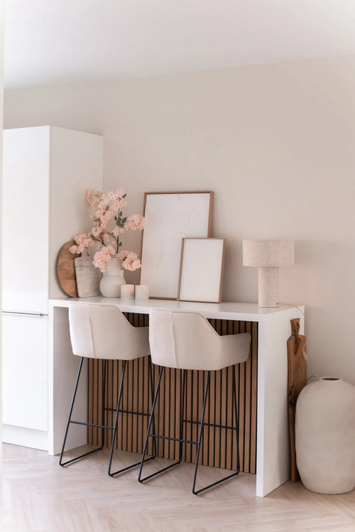 En minimalistisk hemmabar med två beiga pallar med kuddar, en vit disk, inramad konst, en bordslampa, neutraltonad dekoration och Artificiell körsbärsblomma 100 cm ljusrosa i vaser mot en ljus vägg.