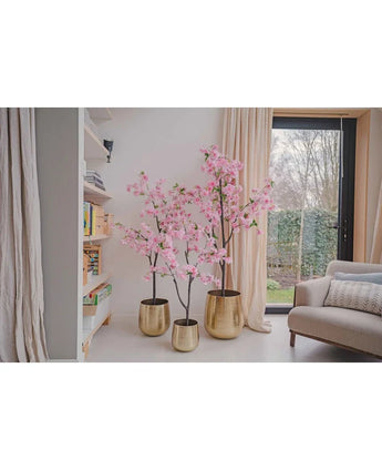 Drie Kunst Kersenbloesem 180 cm roze bomen in gouden potten staan naast een boekenplank en groot raam met gordijnen, naast een lichtgekleurde bank in een moderne, lichte woonkamer.
