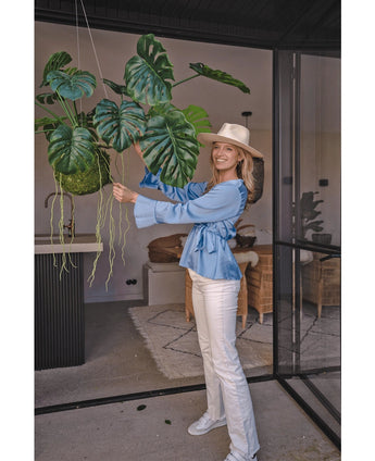 En leende kvinna med hatt, blå blus och vita byxor står inne och håller i en konstgjord hängväxt Monstera Deluxe 60 cm med stora gröna blad, omgiven av modern inredning för en tropisk atmosfär med dekorativa växter.