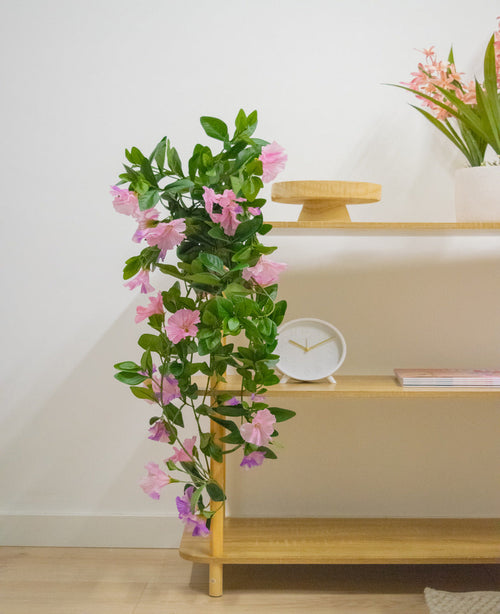En trähylla med konstgjord hängande växt Petunia 70 cm lila, en vit klocka, ett träställ, en bok och en vas med rosa blommor mot en vit vägg.