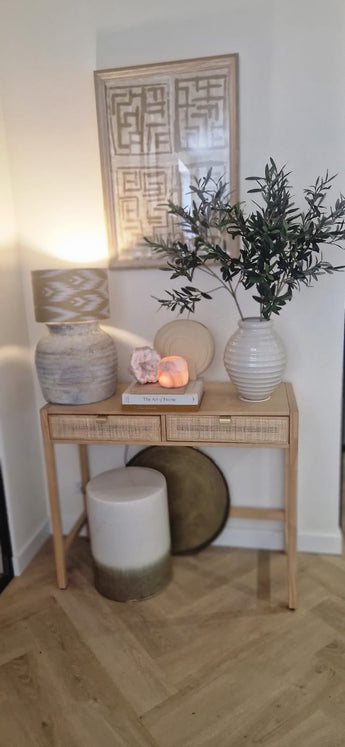 Ett konsolbord i trä med två lådor innehåller en lampa, en stor vit vas med konstgjorda olivblad 90 cm brandskyddsmedel, en mindre vas, böcker och dekorativa stenar. Medelhavsaccenter och inramad abstrakt konst kompletterar utseendet ovan. I botten finns en pall och en rund bricka.