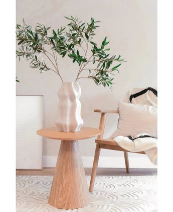 I ett modernt, minimalistiskt vardagsrum med medelhavsdekor står en skulpterad vit vas med det konstgjorda olivbladet 90 cm brandskyddsmedel på ett runt träbord, bredvid en ljus trästol med vita och svarta kuddar.