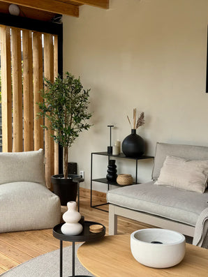 Moderne woonkamer met neutrale tinten, met een lichtgrijze bank, de Kunst olijfboom UV 160 cm potplant, minimalistische zwarte bijzettafeltjes, decoratieve vazen en houten accenten. Natuurlijk licht schijnt door verticale houten lamellen aan de linkerkant.