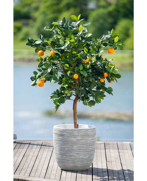 Ett 120 cm konstgjort apelsinträd med mogna frukter står i en vit präglad kruka på en träterrass, med utsikt över ett disigt vattendrag och ett grönt landskap utanför.