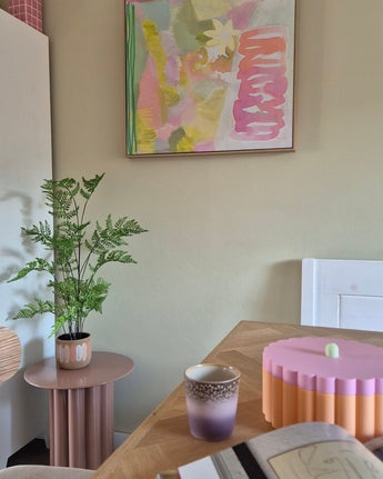 I ett rum med pastelltema finns en 85cm konstgjord ormbunkeväxt på ett rosa sidobord, en lila och guldkopp, en rosa skål med bågad kant, en öppen anteckningsbok och på väggen en abstrakt målning med gröna, rosa och gula toner.