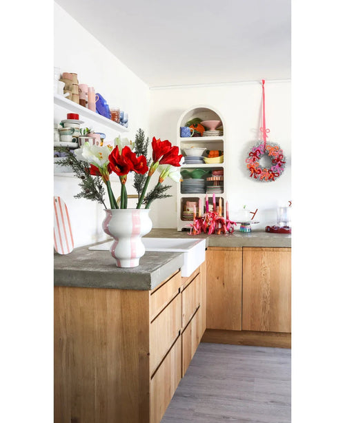 Ett ljust kök med träskåp, öppna vita hyllor med tallrikar och krukor, en festlig krans på väggen och en Konstblomma Amaryllis Real Touch röd 36 cm på bänkskivan i betong.