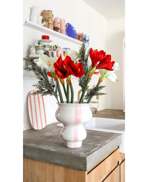 Den konstgjorda blomman Amaryllis Real Touch röd 36 cm står i en vit med rosa randig keramikvas på en bänkskiva i betong i ett ljust kök med hyllor med färgglada porslin i bakgrunden.
