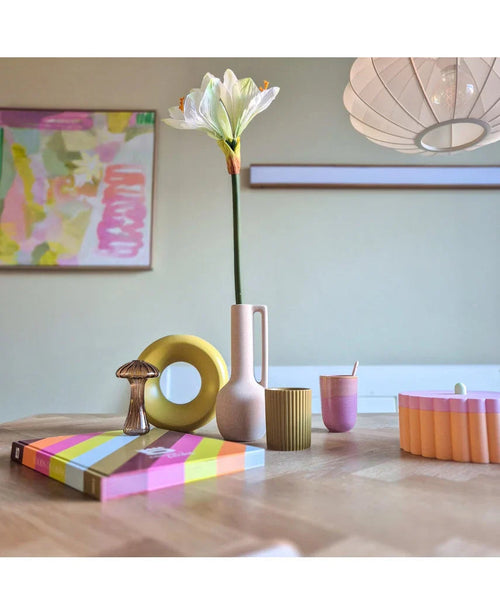 Ett modernt träbord visar färgglada föremål, inklusive en rosa vas innehållande den konstgjorda blomman Amaryllis Real Touch vit 66 cm, en gul ringskulptur, randiga böcker och en rosa kopp. Abstrakt konst accenterar bakgrundsväggen.