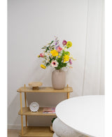 En ljus trähylla visar en beige vas med Artificiell Ranunculus Real Touch Yellow 60cm blommor, en vit klocka och en träbricka. En del av ett runt vitt bord dyker upp i förgrunden mot en vanlig vit vägg.