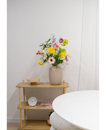 En ljus trähylla visar en beige vas med Artificiell Ranunculus Real Touch Yellow 60cm blommor, en vit klocka och en träbricka. En del av ett runt vitt bord dyker upp i förgrunden mot en vanlig vit vägg.