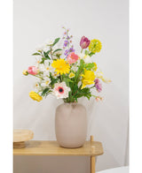 En beige vas med en Artificiell Ranunculus Real Touch Gul 60cm konstgjord blomma står på ett ljust träbord mot en vanlig vit vägg.