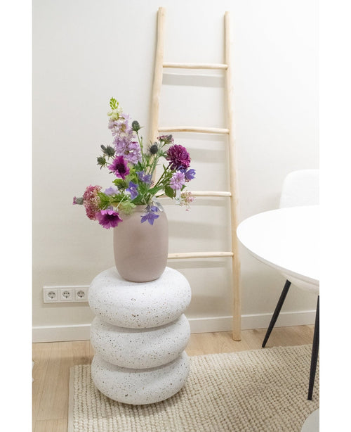 En konstgjord Blomma Harebell av 62 cm lila står i en vas på tre runda, vita stenliknande kuddar. Bakom den står en ljus trästege mot en vit vägg, bredvid ett vitt bord och stol på en beige matta.