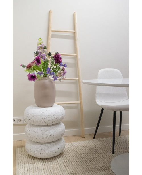 En konstgjord blomma Harebell 62 cm lila står i en beige vas på staplade vita terrazzo pallar. Bakom den finns en stege i ljust trä, nära en vit stol och ett runt bord, allt placerat på en texturerad beige matta i ett minimalistiskt rum.