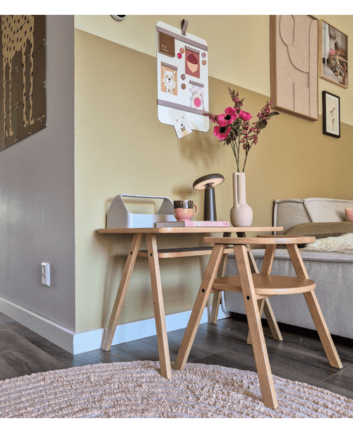 En modern vardagsrumshörna med ett ljust soffbord i trä med konstgjorda blomman Anemone grenar 56 cm rosa i en rosa vas, tillsammans med en lampa och böcker. Bredvid finns en beige soffa, accentuerad av väggkonst och en galleriliknande fototavla på den gula väggen.