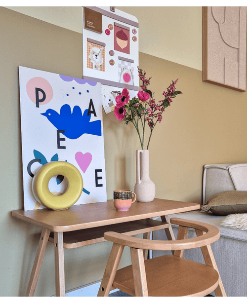 Ett träskrivbord med en matchande stol visar ett blått fågeltryck, konstgjord blomma Anemone grenad 56 cm rosa, en rund gul prydnad och en liten kopp. På övervåningen hänger en anslagstavla med foton och lappar bredvid en beige soffa.