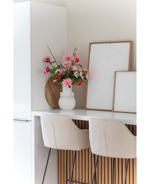 Ett modernt vitt kök med två beiga barstolar, en Artificiell Blomma Dahlia 68 cm rosa i en vit vas på bänken och två inramade abstrakta konstverk mot väggen.