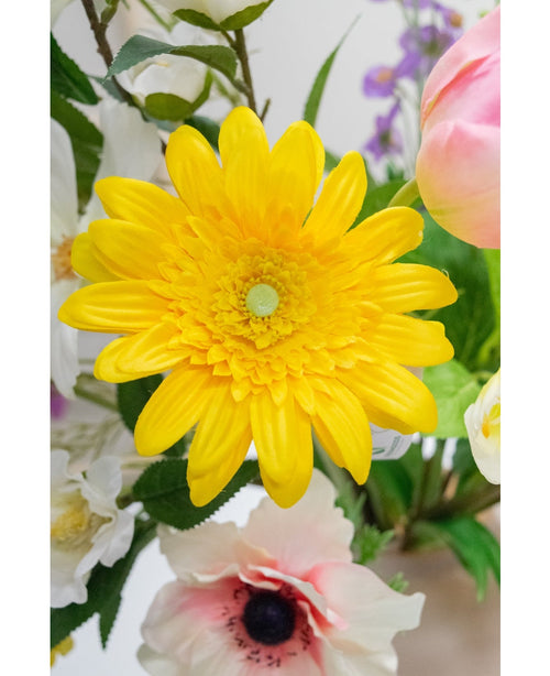 Den konstgjorda blomman Gerbera 53 cm gul, en levande gul konstgjord blomma med lagerblad, står bland andra färgglada konstgjorda blommor och gröna blad, med vita och rosa blommor i bakgrunden.