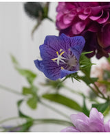 En närbild av den konstgjorda blomman Harebell 62 cm lila, en delikat lila konstgjord blomma med djupa ådror och gula ståndare, mellan gröna blad och mjukt svagt rosa och lila blommor i bakgrunden.