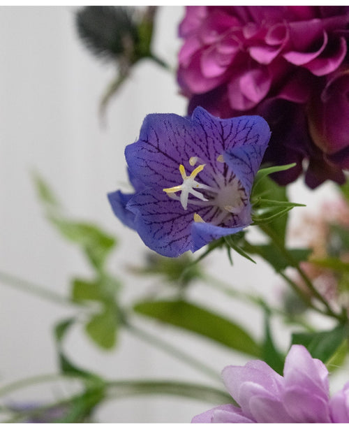 En närbild av den konstgjorda blomman Harebell 62 cm lila, en delikat lila konstgjord blomma med djupa ådror och gula ståndare, mellan gröna blad och mjukt svagt rosa och lila blommor i bakgrunden.